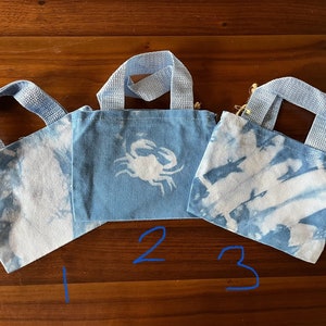 May include: Three small blue tote bags with white tie-dye patterns. The middle bag has a white crab design. The bags have fabric handles.