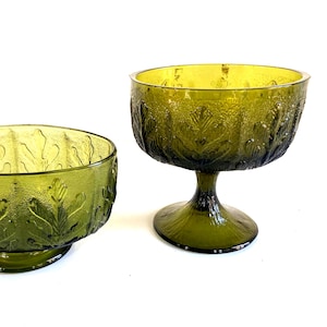 May include: Two green glass bowls with leaf patterns. One is a low bowl, and the other is a stemmed dessert bowl. Both have a textured surface and are set against a white background. The bowls are likely vintage or retro.