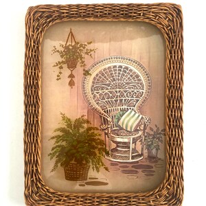 May include: Rectangular woven wicker picture frame. The framed image depicts a peacock chair with a striped pillow, potted plants, and a hanging basket. The background is a light brown color.