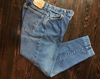 May include: Blue denim jeans with a classic five-pocket design. The jeans have a faded wash and a leather patch on the back. The jeans are folded on a dark wood surface.