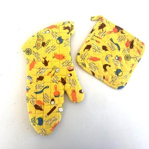 May include: A yellow quilted oven mitt and matching pot holder set. The set features a repeating pattern of various kitchen-themed illustrations in orange, brown, and blue. The oven mitt has a thumb and four finger slots.