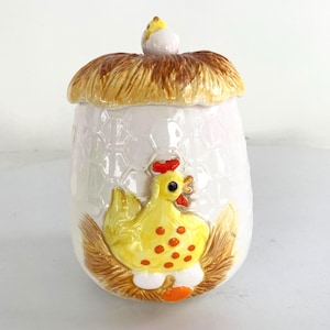 Op de afbeelding: Een keramische eivormige pot met een deksel, met een gele kip met rode stippen en een rode kam. Het deksel is ontworpen om op een nest te lijken met een kleine kuiken erbovenop. De pot is wit met een honingraatpatroon.
