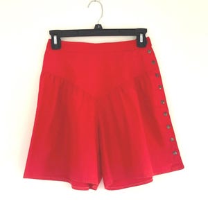 May include: Vibrant red shorts with a high waist and button detailing down the side. The shorts are displayed on a black hanger against a white backdrop. The shorts have a distinctive design.