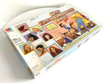 May include: The Baby-Sitters Club board game box. The box features images of the characters and the game's title. The box is rectangular and has the MB Milton Bradley logo. The game is for ages 8 and up, for 2 to 4 players.