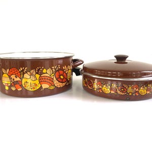 May include: Two-piece brown enamelware cookware set. The set includes a large pot with handles and a smaller oval-shaped casserole dish with a lid. Both pieces feature a decorative border of colorful, stylized fruit and mushroom designs.