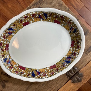 May include: A white oval serving platter with a decorative border of gold, red, and blue mosaic tiles. The border features a repeating pattern of fruit and floral motifs.