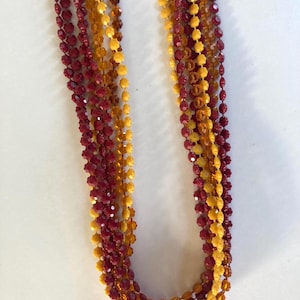 May include: A collection of beaded necklaces in shades of burgundy and amber. The necklaces are made of small, faceted beads strung together, creating a layered effect. The beads have a glossy finish, reflecting light. The necklaces are displayed against a white background.