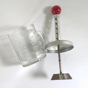 May include: A clear glass measuring cup with embossed markings for 1/4, 1/2, and 3/4 cups alongside a metal hand-operated kitchen tool featuring a red spherical knob, a perforated cylindrical shaft, a circular lid, and a spring-loaded blade base.