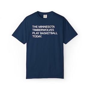 May include: Navy blue t-shirt with the text "THE MINNESOTA TIMBERWOLVES PLAY BASKETBALL TODAY." The shirt is a short-sleeved crew neck style. The text is in a clean, sans-serif font.