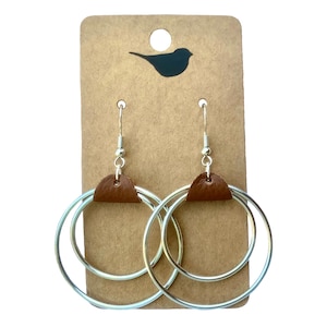 May include: A pair of silver double hoop earrings with brown leather accents. The earrings are hanging on a brown card with a bird silhouette.