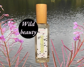 May include: A rectangular perfume bottle with a light brown wooden cap. The label reads "AKINA parfum" with a flock of birds and the words "Wild beauty". The background features a lake and pink wildflowers.