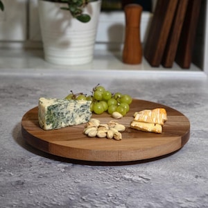 Puede incluir: Una tabla de servir de madera con una cuña de queso azul, uvas verdes, pistachos y algunas rebanadas de queso en la parte superior.