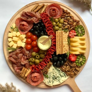 K&ouml;nnte beinhalten: Ein rundes Holz-Charcuterie-Brett, das mit einer Auswahl an Lebensmitteln belegt ist. Das Brett enth&auml;lt Salami-Rosen, K&auml;se, Oliven, Cracker, N&uuml;sse und Obst. Eine kleine Sch&uuml;ssel mit Sauce befindet sich in der Mitte. Das Brett liegt auf einer wei&szlig;en Oberfl&auml;che.