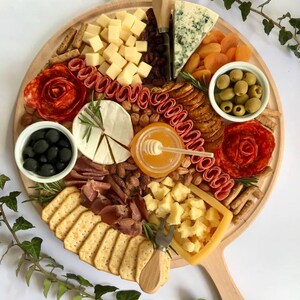 K&ouml;nnte beinhalten: Ein runder Holz-Charcuterie-Brett, gef&uuml;llt mit K&auml;se, Crackern, Oliven, getrockneten Aprikosen, N&uuml;ssen, Salamirosen und Honig. Das Brett hat einen Griff und liegt auf einer wei&szlig;en Oberfl&auml;che.