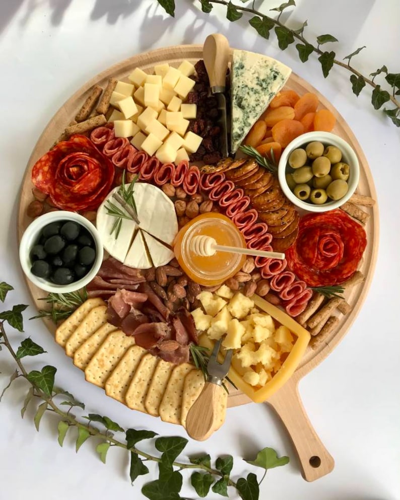K&ouml;nnte beinhalten: Ein runder Holz-Charcuterie-Brett, gef&uuml;llt mit K&auml;se, Crackern, Oliven, getrockneten Aprikosen, N&uuml;ssen, Salamirosen und Honig. Das Brett hat einen Griff und liegt auf einer wei&szlig;en Oberfl&auml;che.