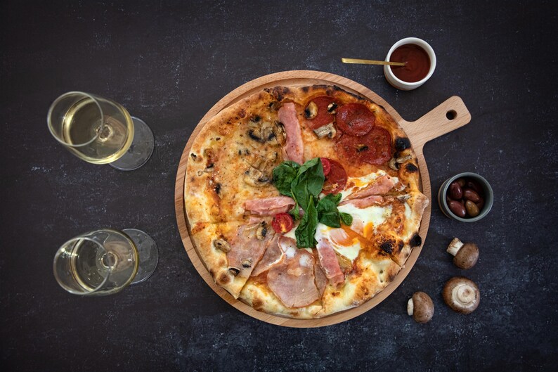 K&ouml;nnte beinhalten: Eine Pizza, unterteilt in Abschnitte mit verschiedenen Bel&auml;gen, darunter Peperoni, Schinken und Pilze, auf einem Holzbrett. Serviert mit Weingl&auml;sern, Oliven, Pilzen und einer kleinen Sch&uuml;ssel mit Sauce. Die Pizza wird auf einer dunklen Oberfl&auml;che pr&auml;sentiert.