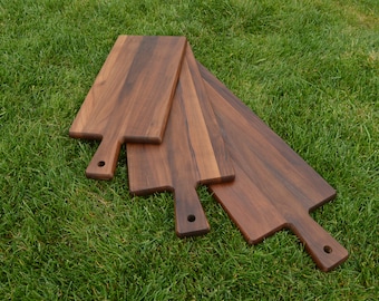 May include: Four wooden chopping boards with handles, made of dark brown walnut wood, arranged in a fan shape on a green grassy surface.
