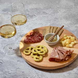 Puede incluir: Una tabla de servir de madera con una variedad de aperitivos, que incluyen salami, queso, galletas, pretzels y miel. Dos vasos de vino blanco están en el fondo.