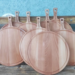 K&ouml;nnte beinhalten: Eine Sammlung runder Pizza-Bretter aus Holz mit Griffen. Jedes Brett hat eine kreisf&ouml;rmige Nut und das Wort "Hometricks" auf dem Griff eingraviert. Die Bretter sind aus hellfarbigem Holz gefertigt und f&auml;cherf&ouml;rmig angeordnet.