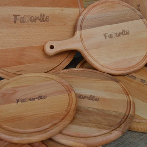K&ouml;nnte beinhalten: Eine Sammlung hellfarbiger Holzschneidebretter mit dem eingravierten Wort "Favorito". Einige Bretter sind rund, andere haben Griffe. Die Bretter haben eine nat&uuml;rliche Holzmaserung und eine glatte Oberfl&auml;che, geeignet f&uuml;r den K&uuml;chengebrauch.