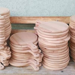K&ouml;nnte beinhalten: Stapel runde Pizzabretter aus Holz mit Griffen. Die Bretter sind hellbraun und scheinen aus unbehandeltem Holz zu bestehen. Sie sind in mehreren Stapeln unterschiedlicher H&ouml;he angeordnet und scheinen zum Verkauf oder zur Verwendung bereit zu sein.
