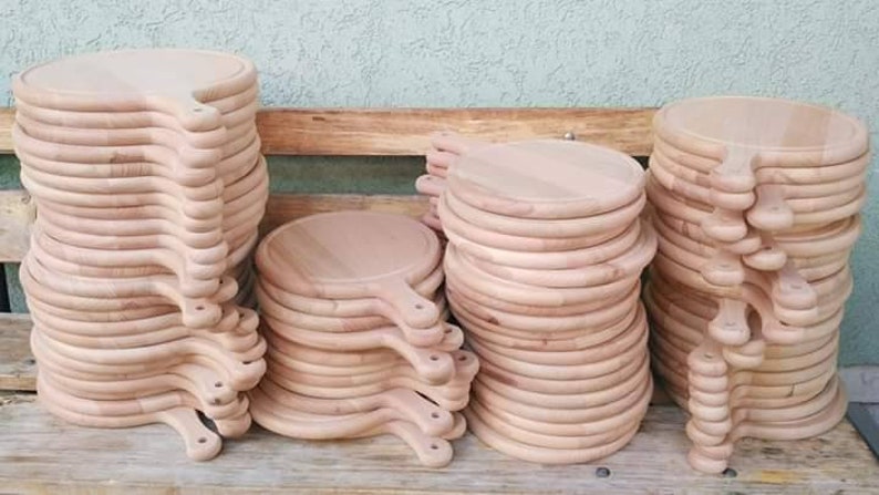 K&ouml;nnte beinhalten: Stapel runde Pizzabretter aus Holz mit Griffen. Die Bretter sind hellbraun und scheinen aus unbehandeltem Holz zu bestehen. Sie sind in mehreren Stapeln unterschiedlicher H&ouml;he angeordnet und scheinen zum Verkauf oder zur Verwendung bereit zu sein.