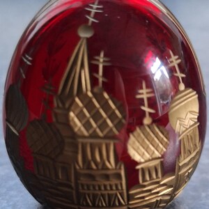 Può includere: Un oggetto decorativo rosso a forma di uovo con disegni architettonici color oro. Il design dorato presenta più cupole e croci, creando un aspetto dettagliato e ornato. L'oggetto ha una finitura lucida.