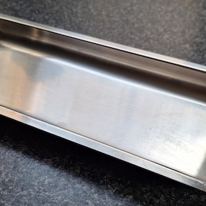 May include: A rectangular stainless steel serving tray with a polished metallic finish and smooth, rounded edges. The tray features two distinct wooden handles attached to each of the shorter ends, providing a contrast to the silver metal surface.