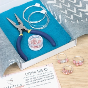 May include: A craft kit for making cocktail rings. The kit includes wire, pliers, beads, and instructions. The kit is packaged in a white box with a pink and blue felt lining. The box also includes a card with the text "Cocktail Ring Kit" and instructions on how to make the rings.