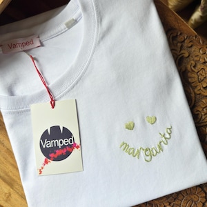 Pode incluir: Camiseta branca com gola redonda e design de carinha sorridente. A carinha sorridente tem olhos em forma de coração e a palavra "margarita" bordada em itálico. Uma etiqueta com a marca "Vamped" está anexada.