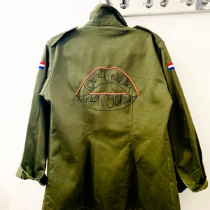 May include: Green military-style jacket with a red, white and blue patch on each sleeve. The back of the jacket has a black embroidered lip with the text "We all need each other" inside.