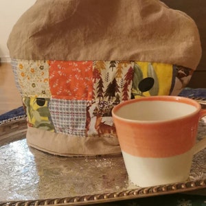 Può includere: Una teiera beige con un design patchwork con motivi floreali, forestali e geometrici. Una tazza da tè bianca e pesca è appoggiata su un vassoio d'argento, creando un'atmosfera accogliente per il tè.