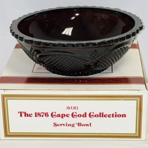 Puede incluir: Un tazón de servir de vidrio rojo oscuro con un borde festoneado y un patrón de diamante. El tazón está en su caja original, que dice "AVON The 1876 Cape Cod Collection Serving Bowl".