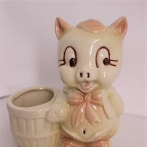 Peut inclure: Un pot en céramique en forme de cochon avec un panier attaché. Le cochon est rose clair avec des yeux bruns et un nœud rose. Le panier est blanc avec un bord brun.