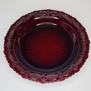 May include: A dark red glass plate with a decorative, scalloped edge. The plate features a textured, starburst pattern in the centre and a detailed border design. The plate is likely vintage or antique, and is suitable for display or serving.
