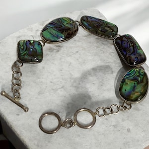May include: A silver chain bracelet with a toggle clasp features five iridescent abalone shell stones in various shapes. The stones have a green, blue, and purple color.