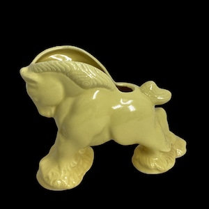 Puede incluir: Un macetero de cerámica amarillo en forma de caballo con un acabado esmaltado. El caballo está de pie sobre sus patas traseras con las patas delanteras levantadas. El macetero tiene un agujero en la parte superior para el drenaje.