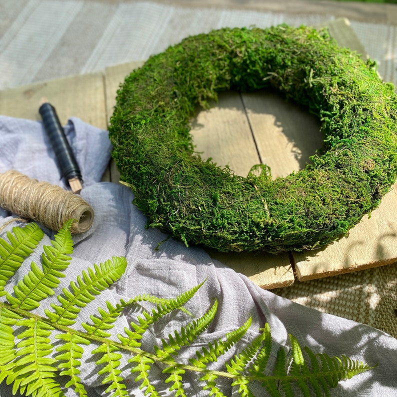 May include: A green moss wreath on a wooden surface with a piece of twine and a fern leaf.
