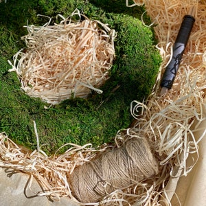 May include: A green moss wreath with a brown twine bow, surrounded by shredded paper in a brown box.