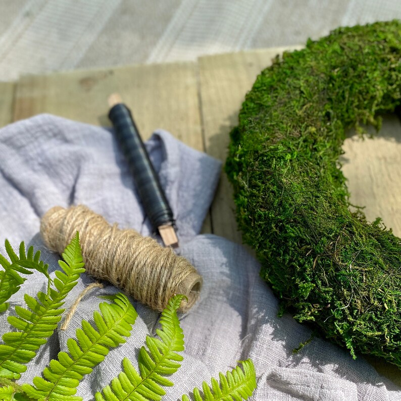 May include: A green moss wreath with a roll of twine and a fern on a gray linen fabric background.