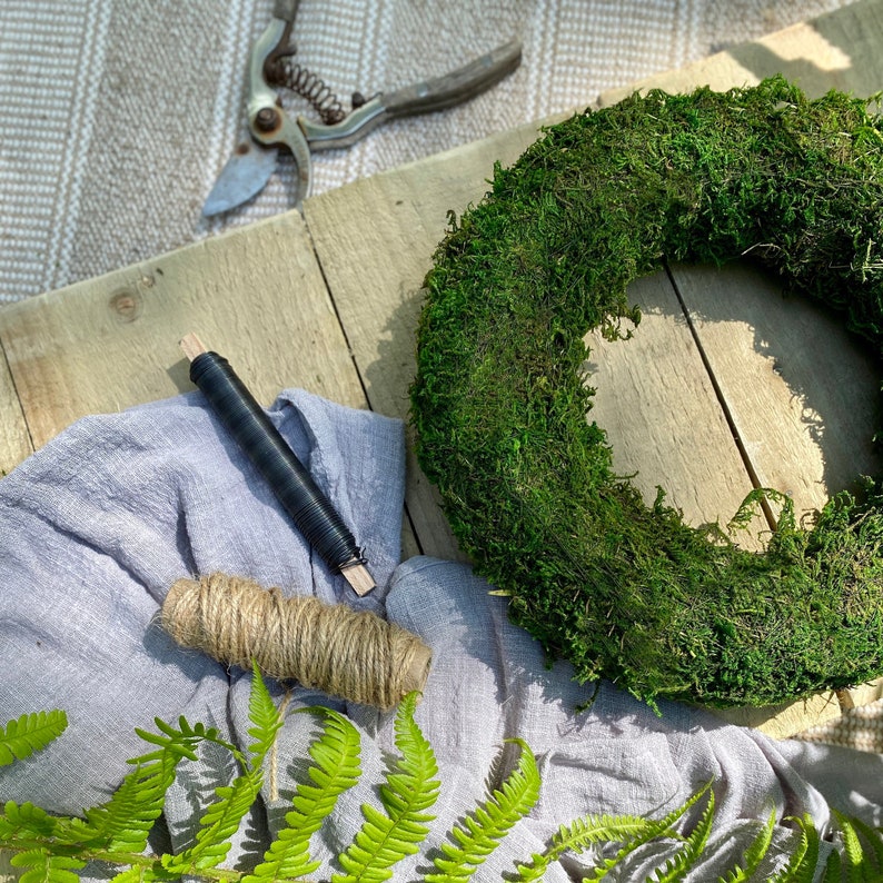 May include: A green moss wreath with a rustic wooden background. The wreath is surrounded by a gray cloth, a spool of twine, a piece of black wire, and green ferns.
