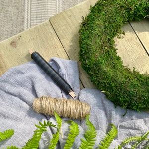 May include: A close-up of a moss wreath, a roll of twine, a piece of black wire, and a piece of gray fabric. The wreath is made of green moss and is lying on a wooden surface. The twine is wrapped around the wire and is being used to create a wreath.