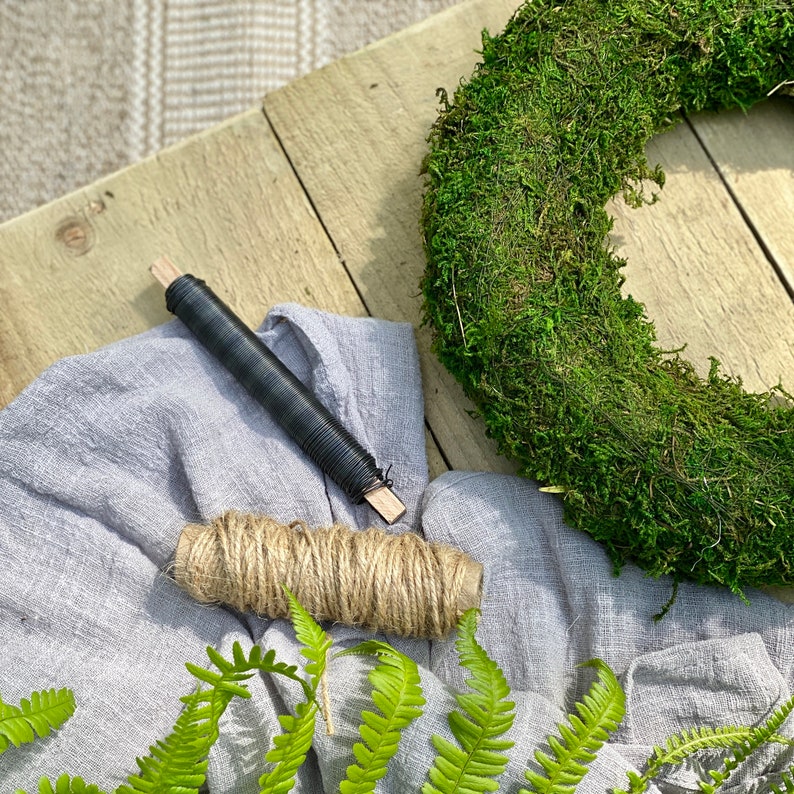 May include: A close-up of a moss wreath, a roll of twine, a piece of black wire, and a piece of gray fabric. The wreath is made of green moss and is lying on a wooden surface. The twine is wrapped around the wire and is being used to create a wreath.