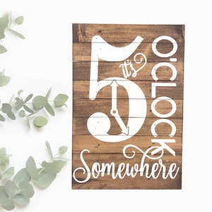 May include: A wooden sign with the text "It's 5 o'clock somewhere" in white paint. The number 5 is styled as a clock face with hands pointing to 5 o'clock.