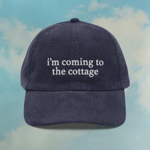 Könnte beinhalten: Marineblaue Baseballkappe aus Cord mit dem weißen, gestickten Text "I'm coming to the cottage". Die Kappe hat einen gebogenen Schirm und eine runde Oberseite. Der Hintergrund ist ein hellblauer Himmel mit weißen Wolken.