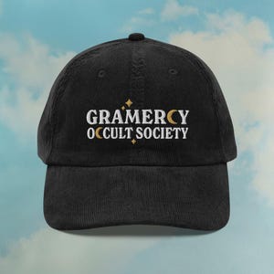 May include: Black corduroy baseball cap with the text "GRAMERCY OCCULT SOCIETY" embroidered in white, with gold star and crescent moon accents. The cap has a curved brim and a top button. The background is a light blue sky with clouds.
