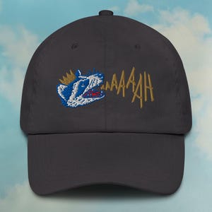 Peut inclure: Une casquette de baseball bleu foncé avec une image brodée d'un animal couronné avec une couronne et le mot "MAAAAH" en fil doré.