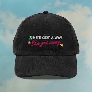 Może przedstawiać: Czarna sztruksowa czapka bejsbolowa z haftowanym tekstem. Tekst brzmi "S HE'S GOT A WAY" w kolorze zielonym i "She got away" w kolorze różowym, z żółtą gwiazdką. Czapka ma zakrzywiony daszek i guzik na górze.