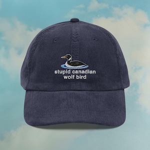 Peut inclure: Casquette de baseball en velours côtelé bleu marine avec un motif brodé d'un oiseau sur l'eau et le texte "stupid canadian wolf bird". La casquette a une visière incurvée et un bouton sur le dessus.