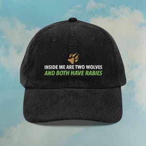 Puede incluir: Gorra de béisbol de pana negra con el texto "INSIDE ME ARE TWO WOLVES AND BOTH HAVE RABIES" en blanco y verde. Un gráfico de huella de pata está encima del texto. La gorra tiene una visera curva.